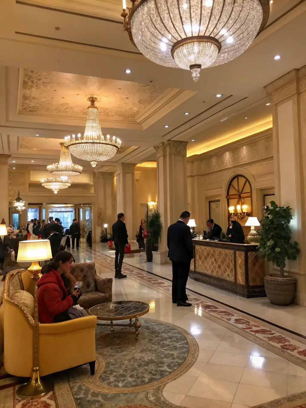 A grand hotel lobby with a sweeping staircase, elegant chandeliers, and a concierge desk. The scene is bustling with guests checking in and out, creating a vibrant atmosphere.